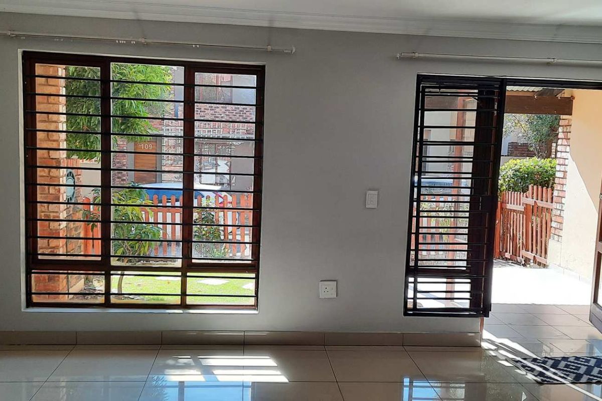 A bright, empty living area featuring tiled floors, large windows with security bars, and direct access to an outdoor patio space.