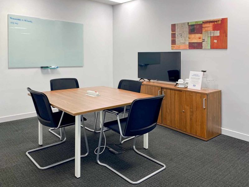 A functional office meeting room featuring a square table, wall-mounted whiteboard, and carpeted flooring, suitable for professional collaboration.