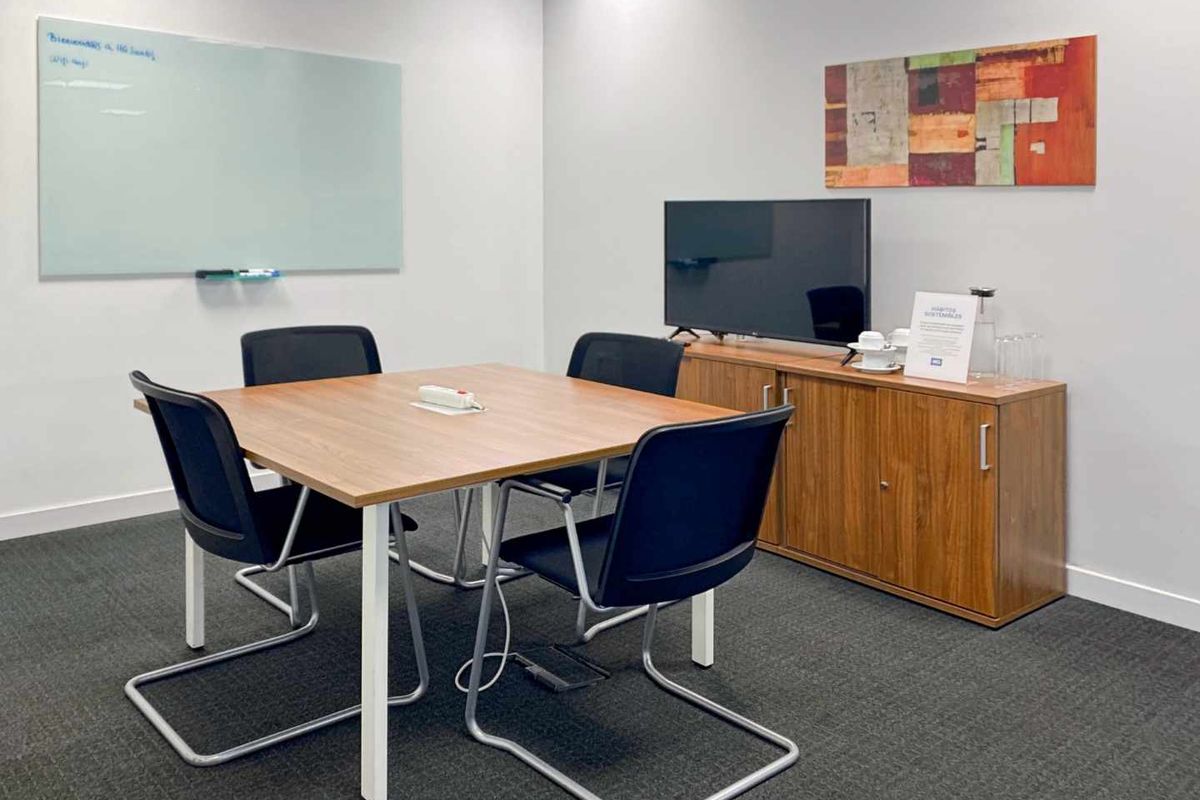 A functional office meeting room featuring a square table, wall-mounted whiteboard, and carpeted flooring, suitable for professional collaboration.