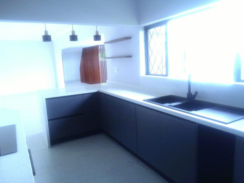 Modern kitchen featuring dark cabinetry, white stone countertops, black sink fixtures, and minimalist floating shelves in a bright, open-plan layout.