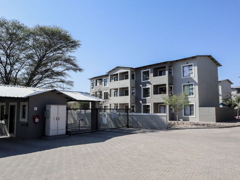 Exterior view of the apartment complex entrance featuring a security gate, guard house, and paved driveway leading to the multi-story residential building.