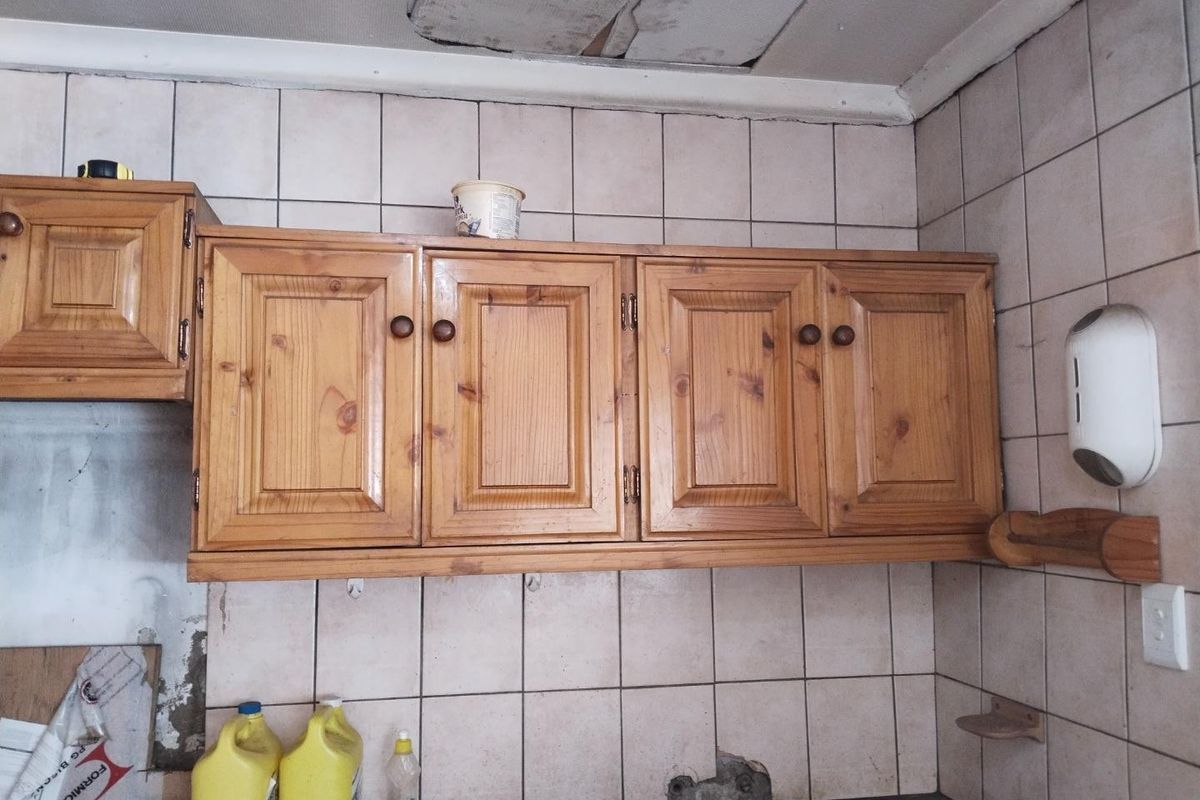 A kitchen area featuring dated wooden wall-mounted cabinetry, tiled splashbacks, and visible damage to the ceiling and wall surfaces.