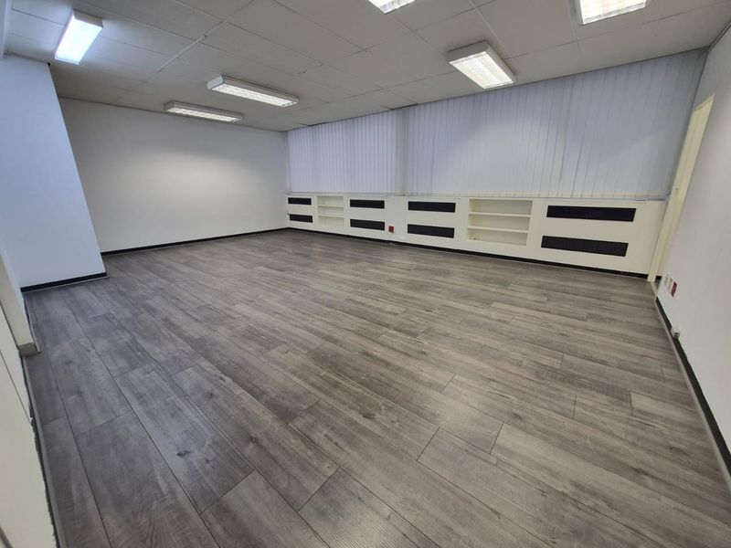A spacious, empty office featuring modern grey laminate flooring, built-in shelving units, vertical blinds, and a suspended ceiling with integrated lighting.