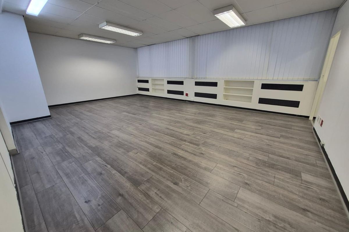 A spacious, empty office featuring modern grey laminate flooring, built-in shelving units, vertical blinds, and a suspended ceiling with integrated lighting.