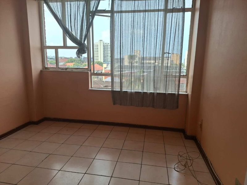 An empty bedroom featuring tiled flooring and a large window with natural light, requiring cosmetic updates to the wall finishes.