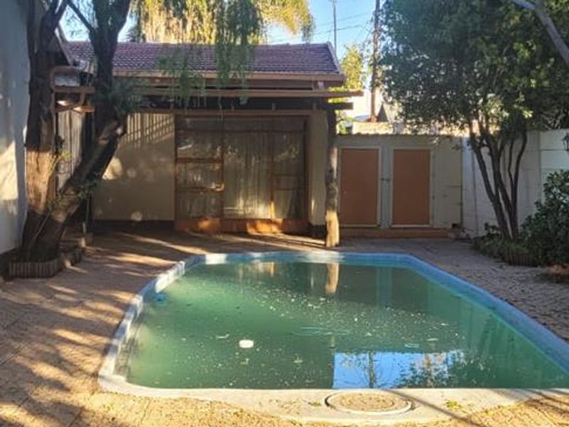 Outdoor pool area featuring a paved patio, covered patio space, and established trees, requiring maintenance to the pool water and surrounding area.