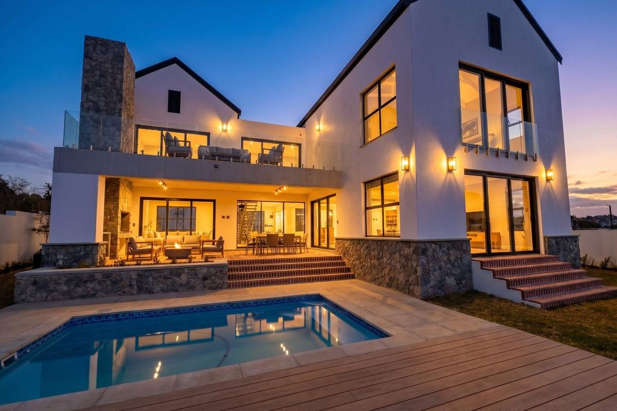 Stunning rear elevation featuring a modern swimming pool, expansive decking, covered patio with built-in fireplace, and elegant stone and glass architectural details.