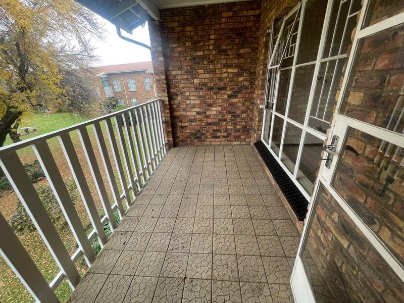 A covered balcony featuring brick walls, tiled flooring, and a metal safety railing, offering an outdoor space with views of the surrounding garden area.