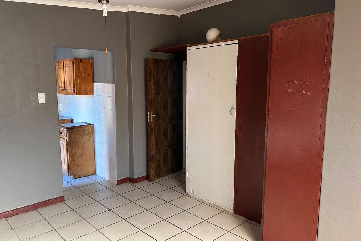 An interior view showing a living area with tiled flooring and built-in storage, leading into a separate kitchen space with wooden cabinetry.