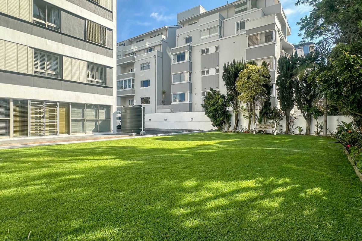 A well-maintained communal garden area featuring a lush green lawn, boundary wall, and water storage tank, situated between apartment building blocks.