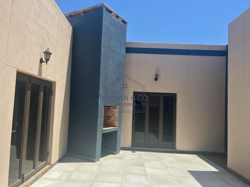 A paved outdoor patio area featuring a built-in braai, modern aluminium glass doors, and exterior wall lighting, providing a clean space for outdoor entertainment.