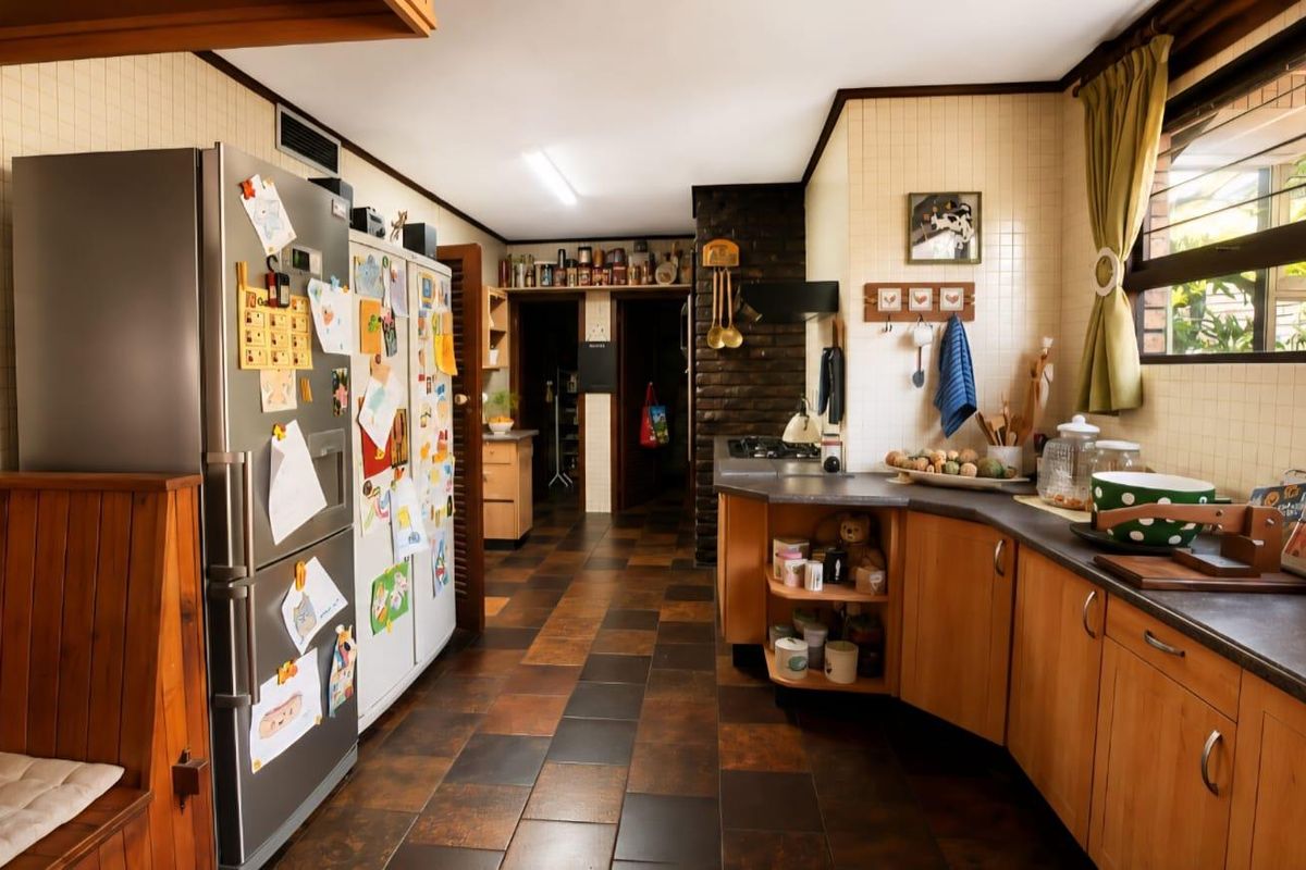 A functional kitchen featuring wooden cabinetry, dark laminate countertops, tiled splashbacks, and a distinctive brick feature wall with durable tiled flooring.