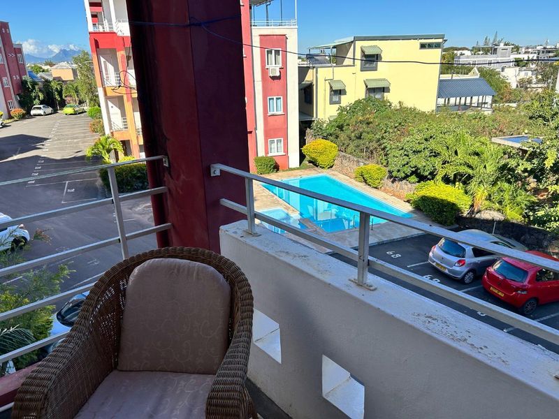 A balcony overlooking the apartment complex parking area and communal swimming pool, featuring a metal balustrade and concrete parapet wall.