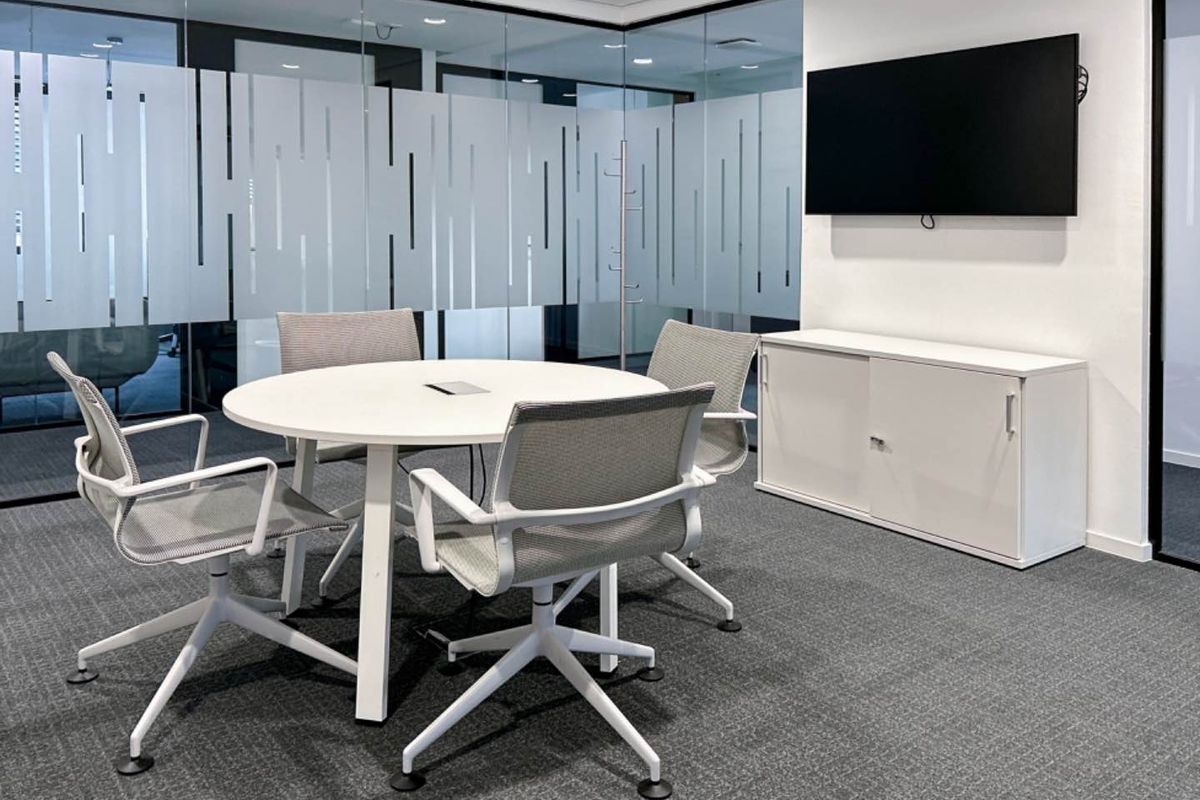 Modern office meeting room featuring glass partition walls with privacy film, carpeted flooring, and a wall-mounted television.