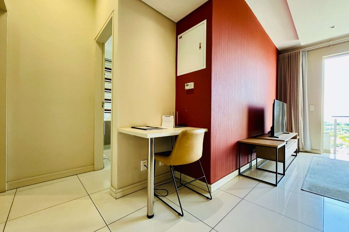 A modern living area featuring tiled flooring, a striking red accent wall, and a built-in desk nook, leading to a bright balcony space.