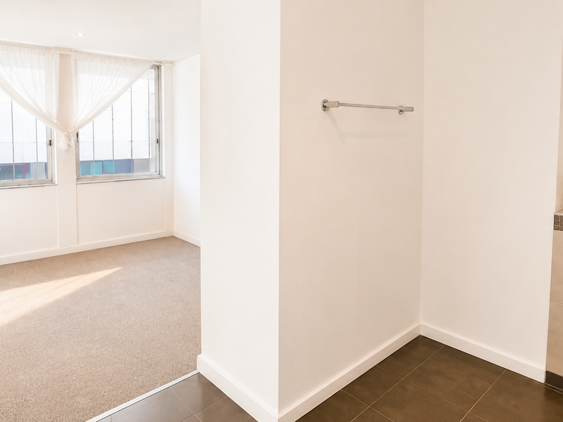A bright, empty bedroom featuring neutral carpeting, an integrated pedestal sink area with tiled splashback, and large windows allowing for natural light.