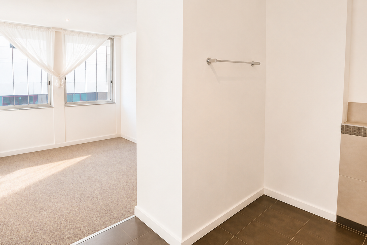 A bright, empty bedroom featuring neutral carpeting, an integrated pedestal sink area with tiled splashback, and large windows allowing for natural light.