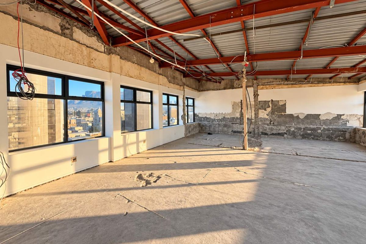 An expansive open-plan penthouse space in shell condition, featuring exposed steel roof trusses, concrete flooring, and large windows with city views.