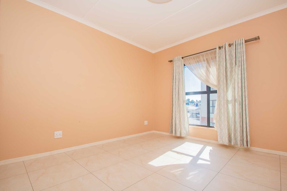 A bright, empty bedroom featuring neutral tiled flooring, peach-toned walls, and a large window with installed curtain rod.