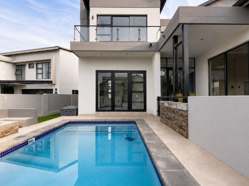 Modern double-story rear elevation featuring a swimming pool, stone-clad feature wall, and a glass-balustrade balcony overlooking the patio area.