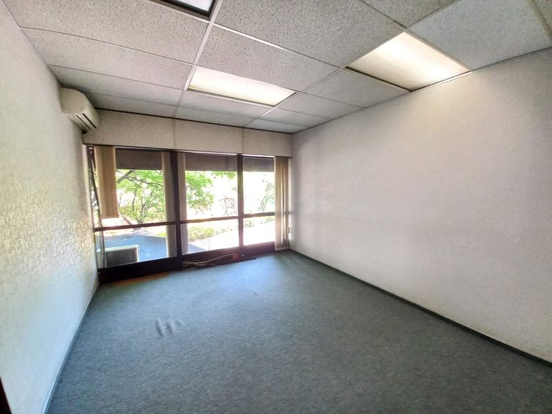 A vacant office space featuring a drop ceiling with recessed lighting, wall-mounted air conditioning unit, and large windows providing natural light.