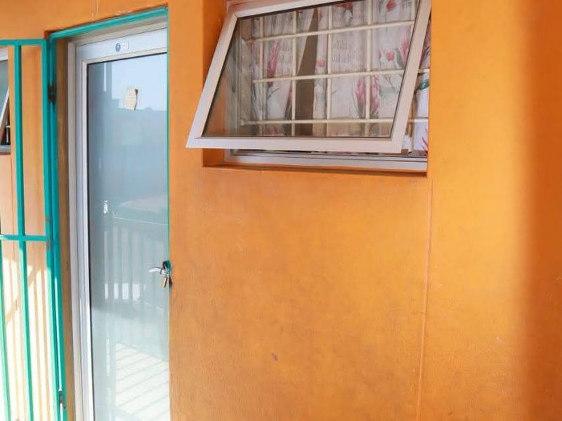 A covered balcony area featuring a security gate, glass door, and top-hung window with burglar bars, finished with tiled flooring and exterior wall lighting.