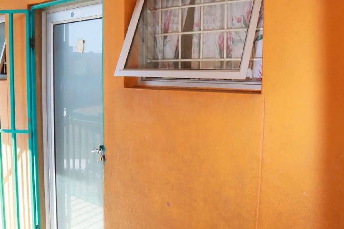 A covered balcony area featuring a security gate, glass door, and top-hung window with burglar bars, finished with tiled flooring and exterior wall lighting.