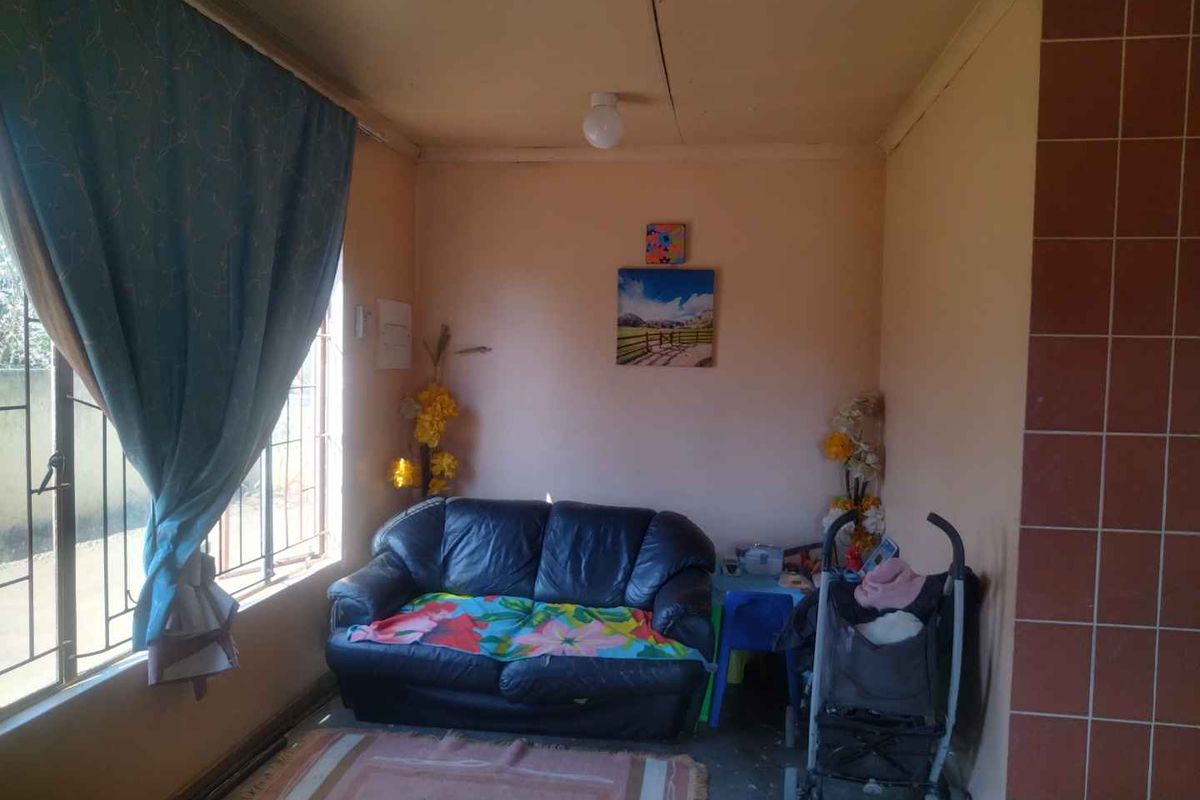 A compact living area featuring a large window with security bars, tiled wall accents, and basic ceiling lighting.