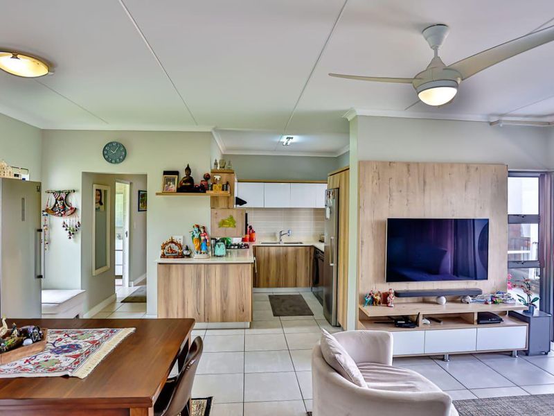 An open-plan living and kitchen area featuring modern wood-finish cabinetry, tiled flooring, a ceiling fan, and a built-in media unit with balcony access.