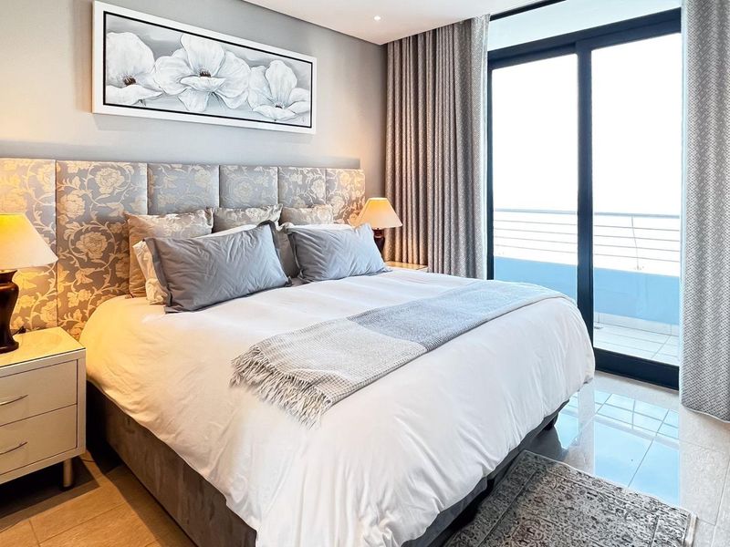 A well-appointed bedroom featuring a large upholstered headboard, tiled flooring, and direct access to a private balcony through sliding glass doors.