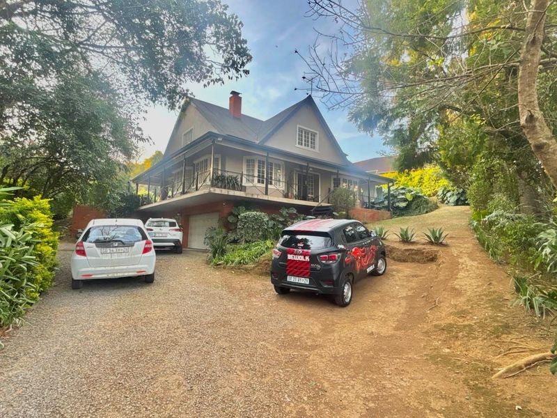 A charming double-story house featuring a wrap-around veranda, pitched roof, and a gravel driveway set within a lush, mature garden.