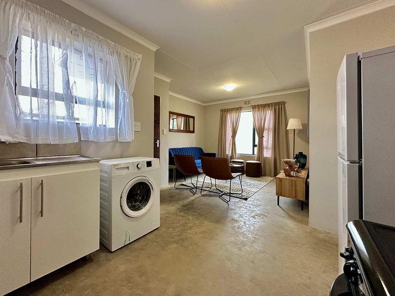 Open-plan living area featuring a compact kitchen section with built-in cabinetry and laundry plumbing, flowing into a combined lounge space with concrete flooring.