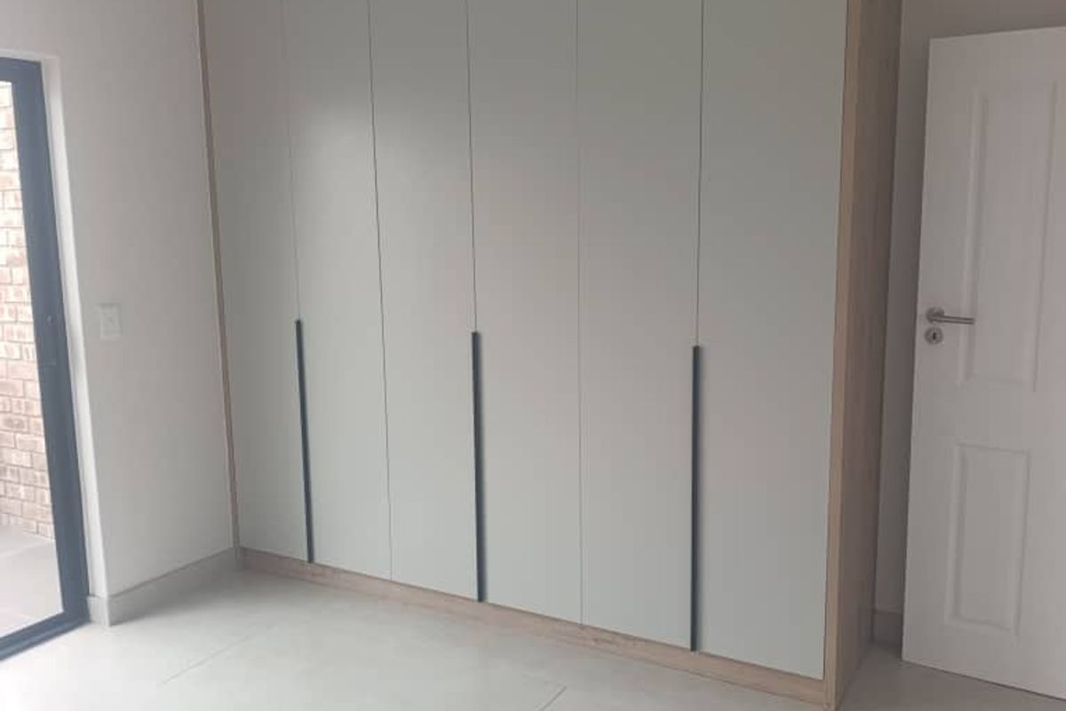 A bright, modern bedroom featuring neutral tiled flooring and extensive floor-to-ceiling built-in cupboards with contemporary hardware.