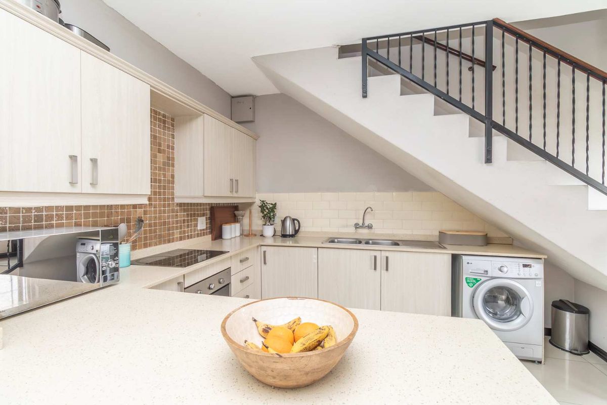 Compact kitchen featuring light-toned cabinetry, stone-look countertops, tiled backsplash, built-in oven, hob, and integrated space for a washing machine under the staircase.