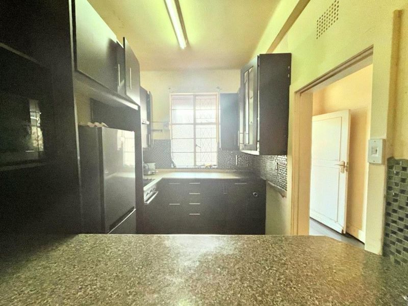 A functional kitchen featuring dark built-in cabinetry, mosaic tile backsplashes, and laminate countertops, illuminated by a central fluorescent light fixture.
