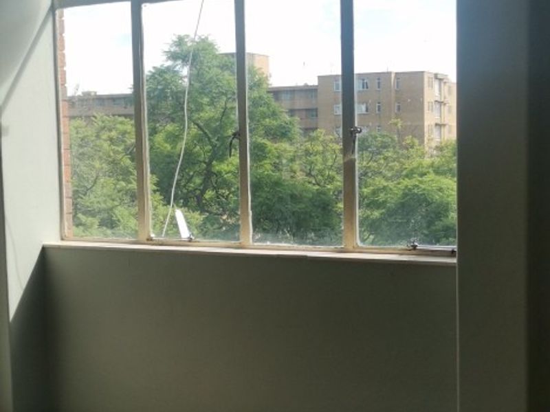 An empty living area featuring large metal-framed windows with an urban view and damaged floor tiling in need of repair.