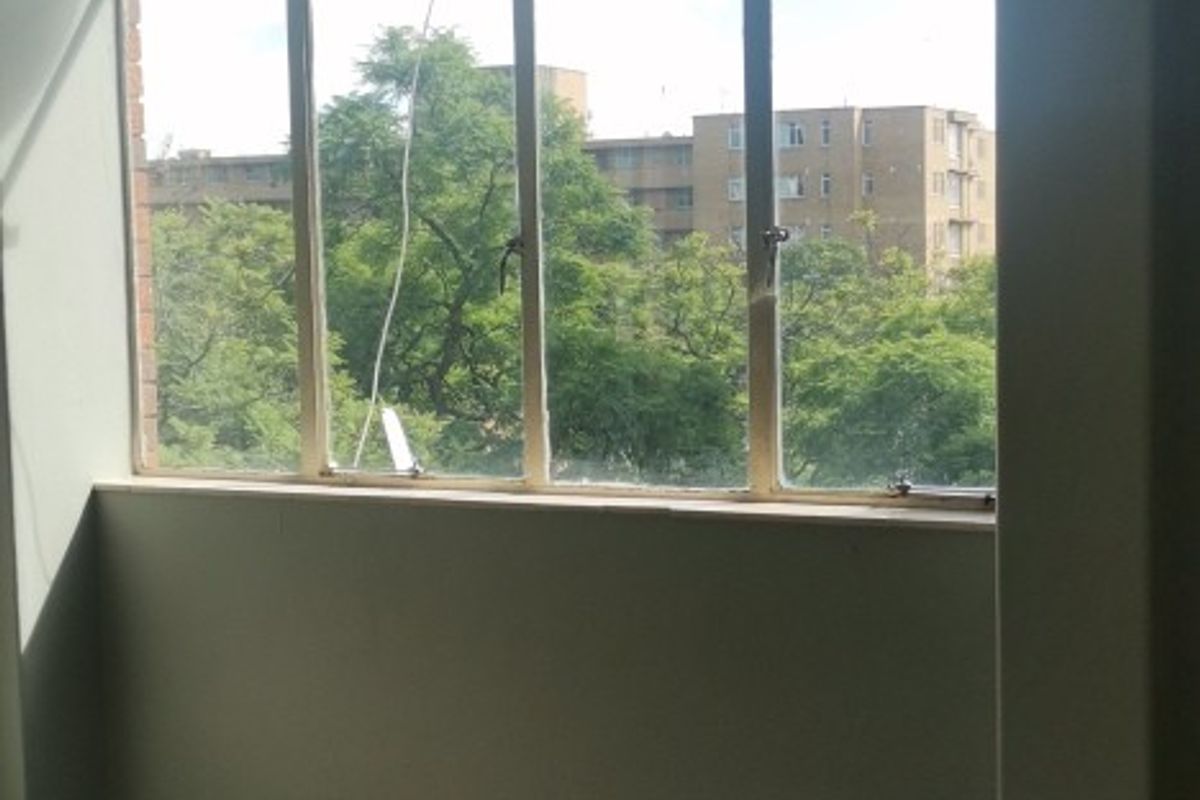 An empty living area featuring large metal-framed windows with an urban view and damaged floor tiling in need of repair.