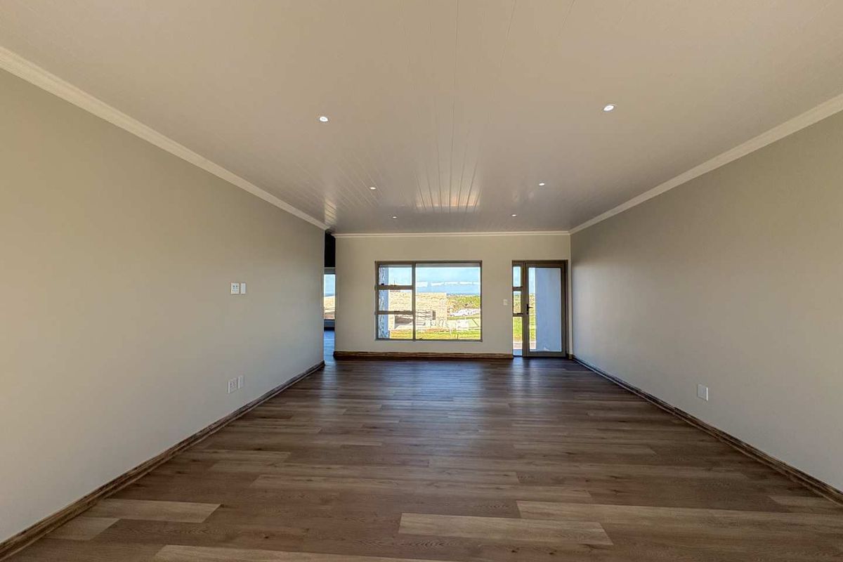 A spacious, empty living area featuring modern wood-look flooring, recessed ceiling lighting, and a large window providing natural light and exterior views.