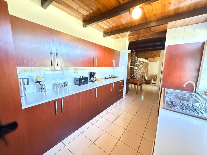 A functional kitchen featuring warm wooden cabinetry, tiled flooring, and exposed timber ceiling beams, leading into an adjacent dining area with a stone feature wall.