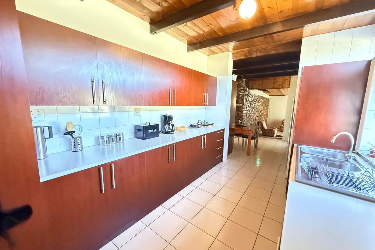 A functional kitchen featuring warm wooden cabinetry, tiled flooring, and exposed timber ceiling beams, leading into an adjacent dining area with a stone feature wall.