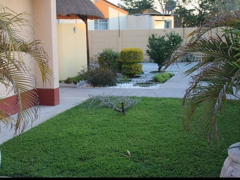 A well-maintained rear garden area featuring a paved walkway, landscaped garden beds, and a thatched roof structure against a secure boundary wall.