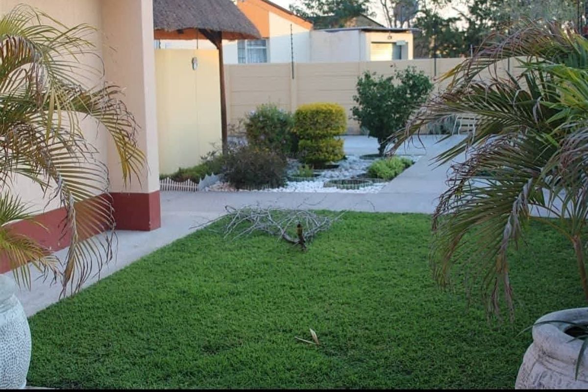 A well-maintained rear garden area featuring a paved walkway, landscaped garden beds, and a thatched roof structure against a secure boundary wall.