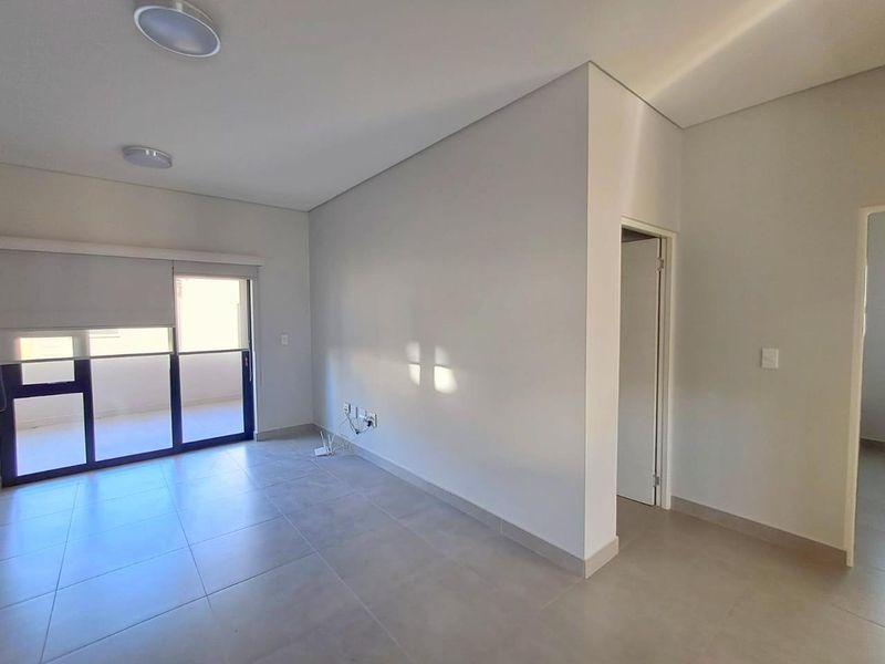 A bright, empty living area featuring neutral tiled flooring, modern ceiling lighting, and a sliding glass door providing access to an outdoor balcony.
