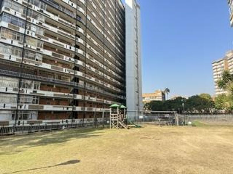View of the apartment building exterior featuring a communal garden area with playground equipment and perimeter fencing.