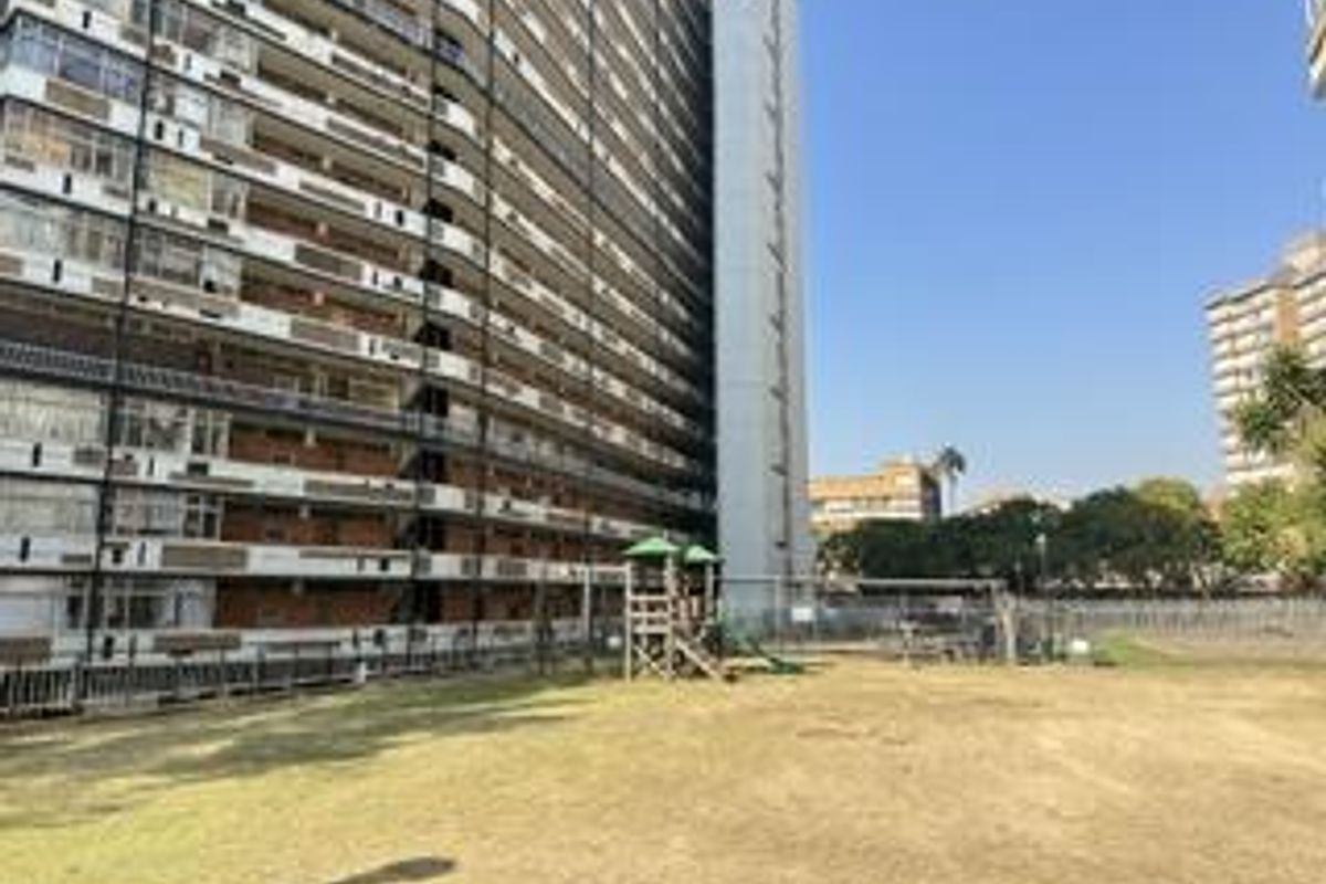 View of the apartment building exterior featuring a communal garden area with playground equipment and perimeter fencing.