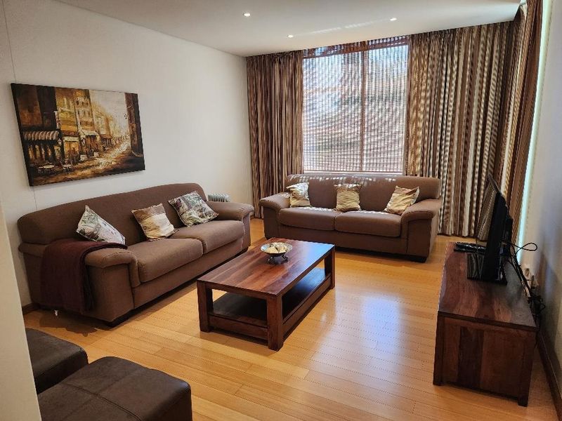 A comfortable living room featuring warm timber flooring, recessed downlighting, and a large window with floor-to-ceiling curtains.