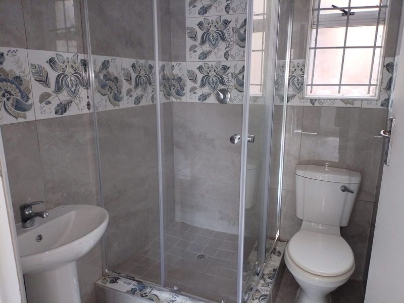 A clean, functional bathroom featuring a glass-enclosed shower, pedestal washbasin, and toilet, finished with decorative tiled walls and a window with security bars.