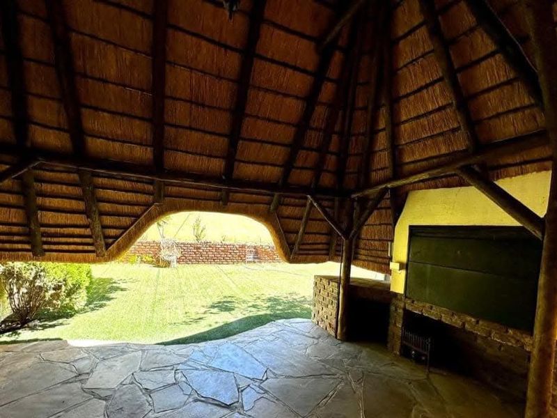 Covered outdoor patio featuring a traditional thatched roof, exposed timber structure, built-in braai, and stone flooring overlooking the garden.
