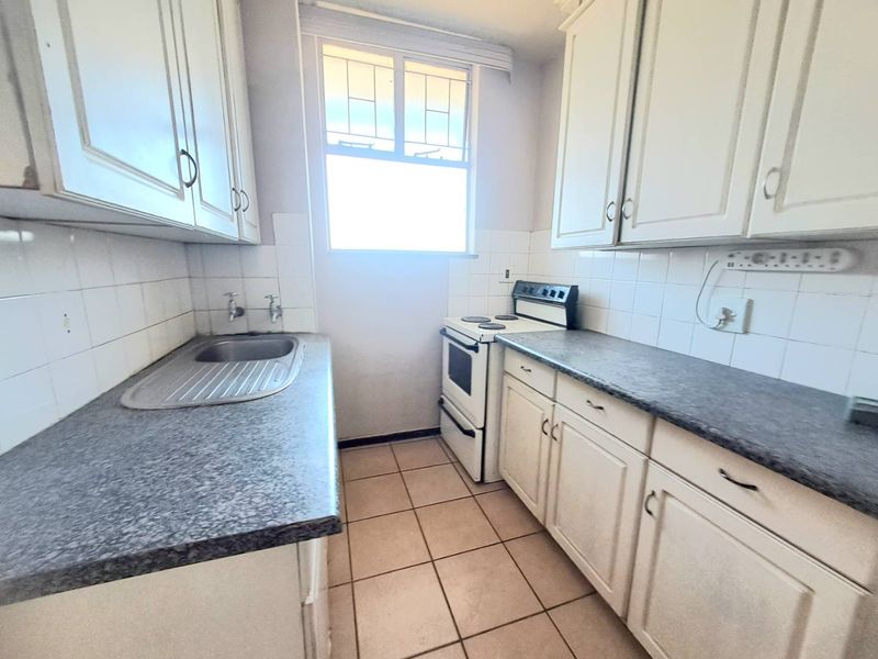 A functional galley-style kitchen featuring white cabinetry, laminate countertops, a stainless steel sink, and a freestanding stove with tiled flooring and backsplash.