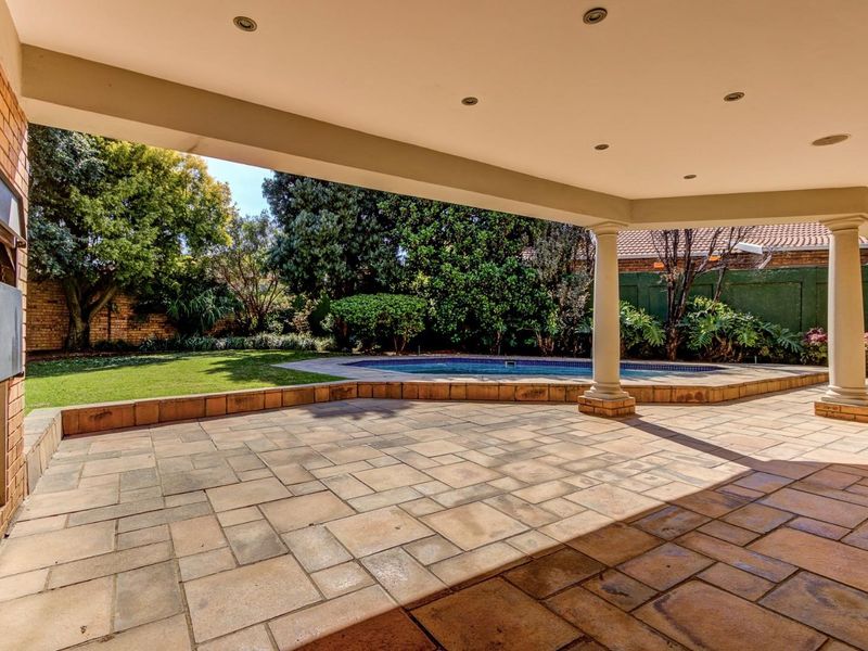 Spacious covered patio featuring paved flooring, a built-in braai, and recessed lighting, overlooking a private swimming pool and landscaped garden area.